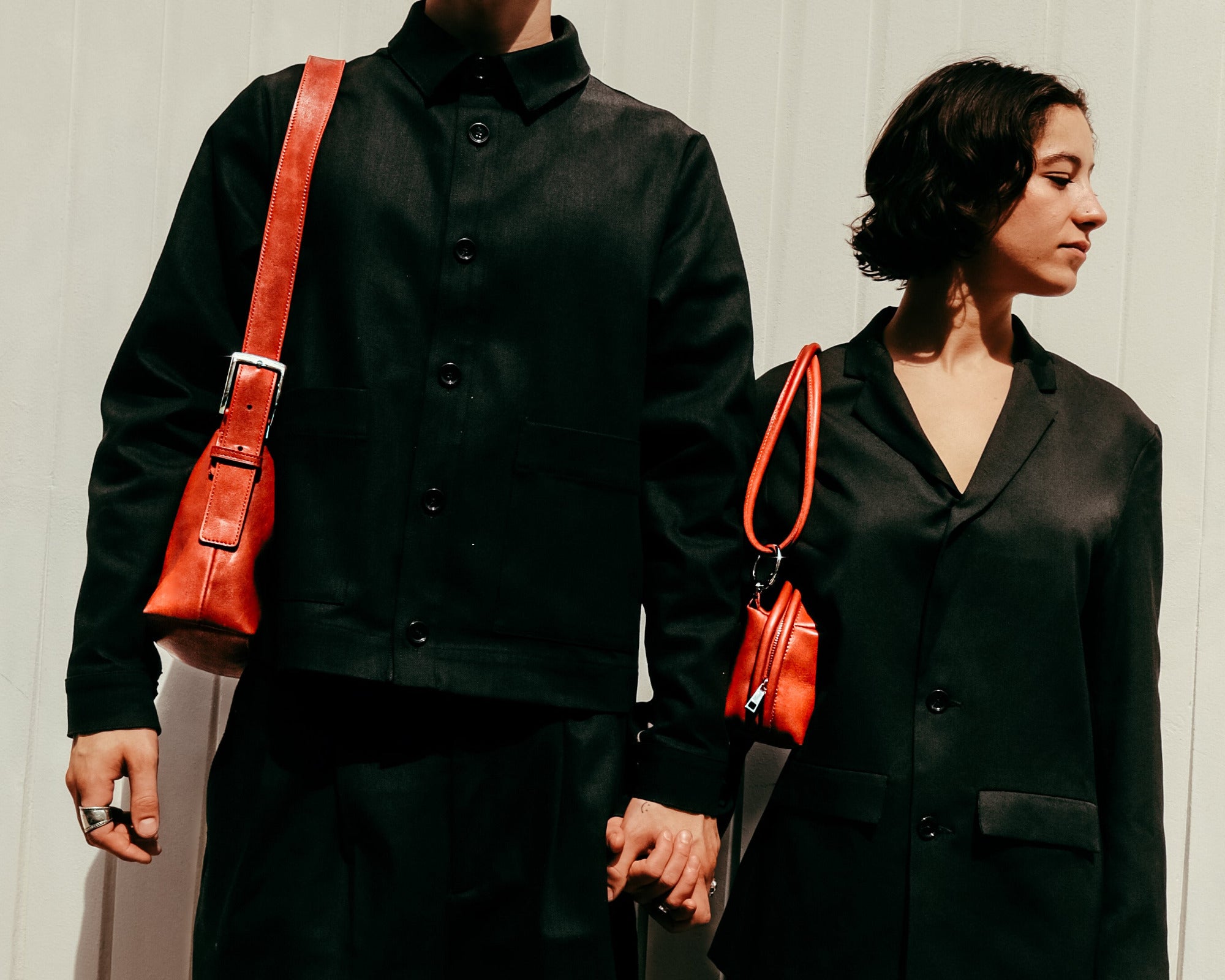 Two people in black outfits with red handbags against a neutral background