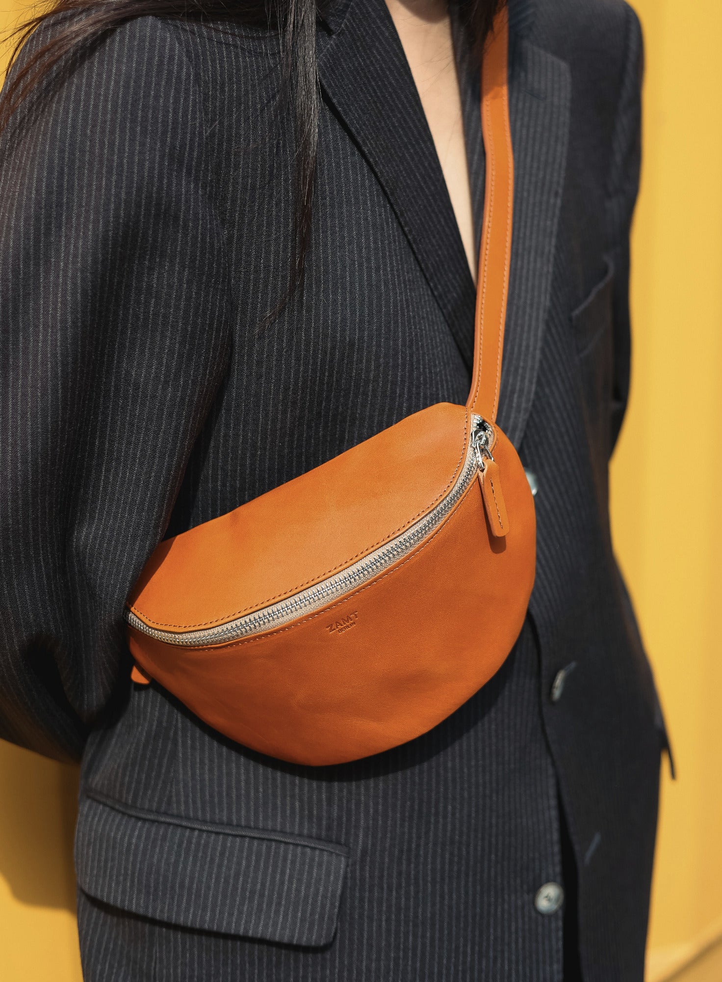 Person wearing a dark pinstripe suit with an orange crossbody bag against a yellow background